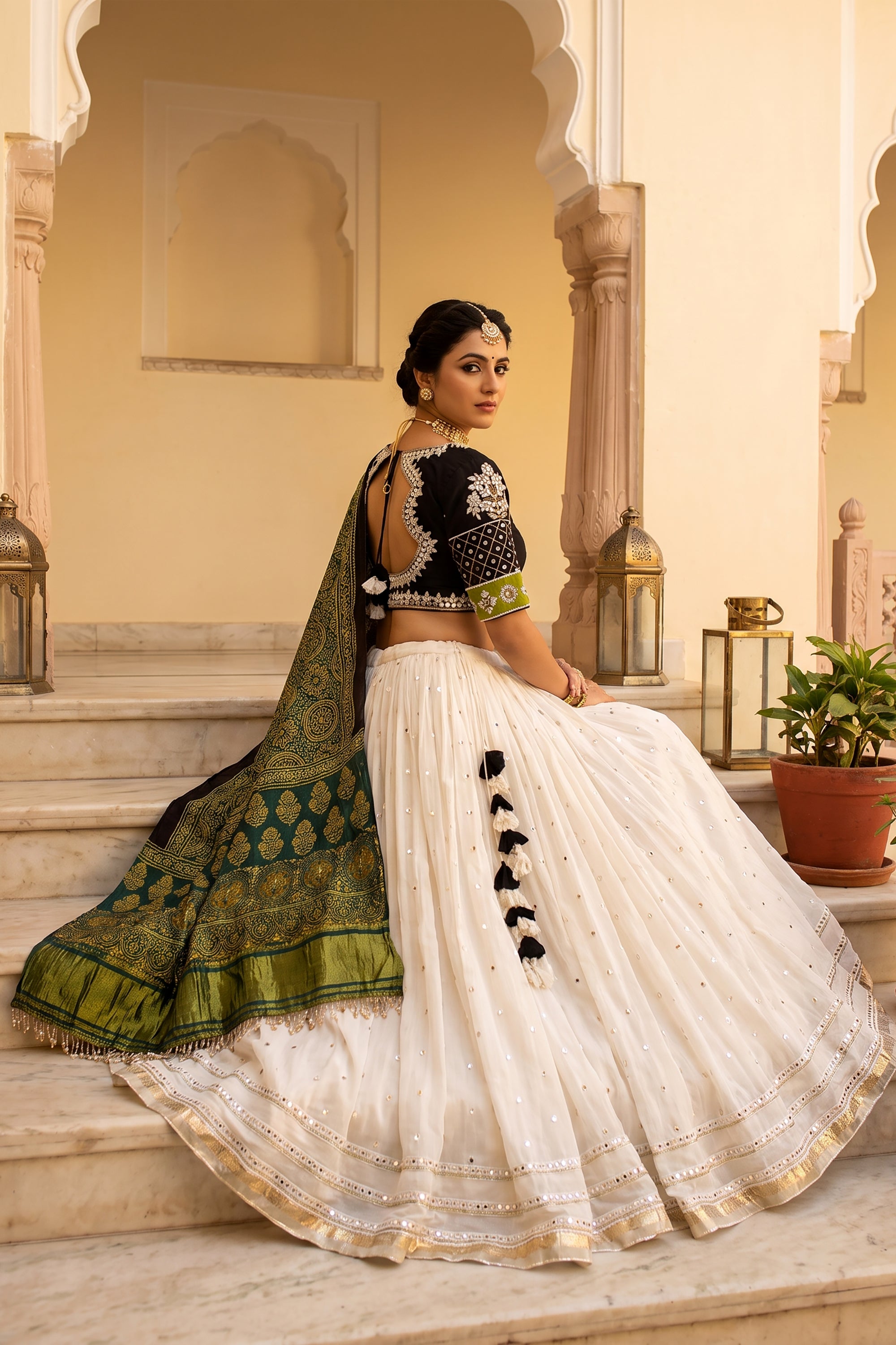 A woman smiling and twirling in a white flared georgette chaniya choli with subtle mirror work at the hem. The outfit is paired with a black V-neck blouse featuring embroidered sleeves and a draped green Ajrakh print dupatta.