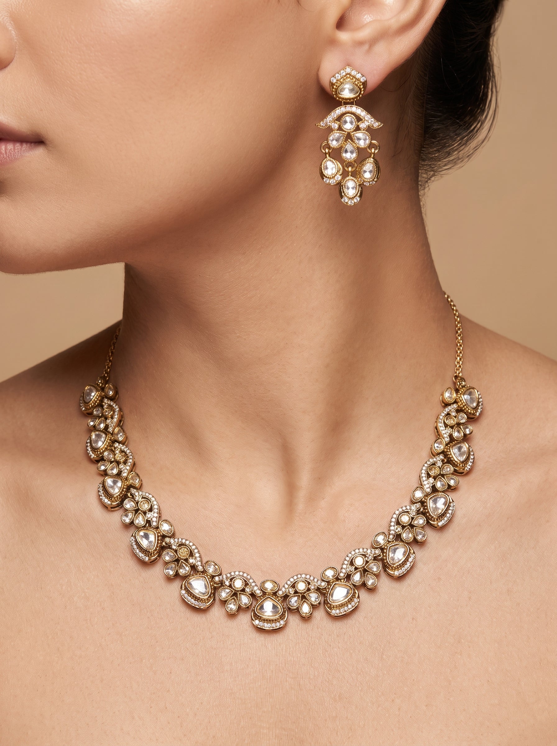Gold-toned statement jewelry set featuring a wide collar necklace and a matching drop earring, embellished with clear stones and pearls, shown on a model against a warm beige background.