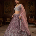 A model wears a mauve raw silk lehenga choli decorated with pastel pink and aqua Resham embroidery, featuring a draped sheer net dupatta in a luxurious mansion setting.