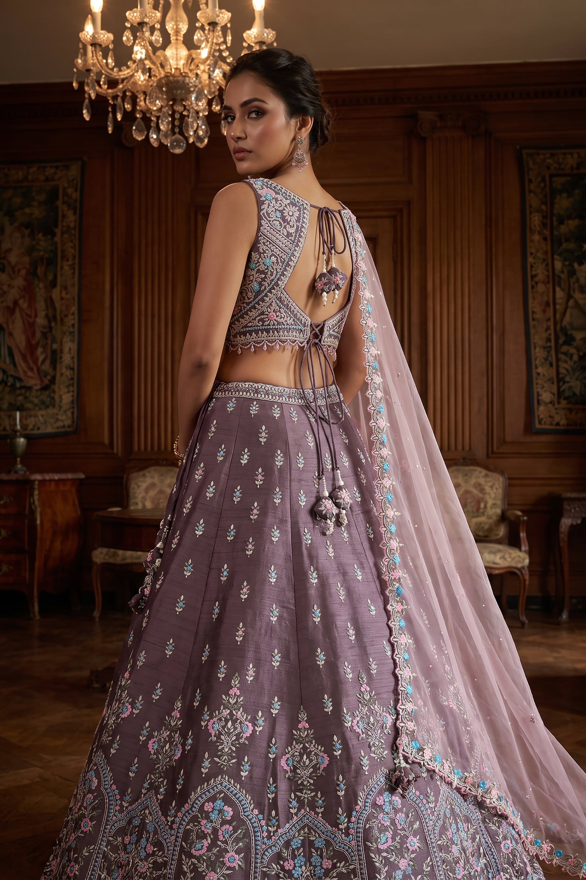 A model wears a mauve raw silk lehenga choli decorated with pastel pink and aqua Resham embroidery, featuring a draped sheer net dupatta in a luxurious mansion setting.