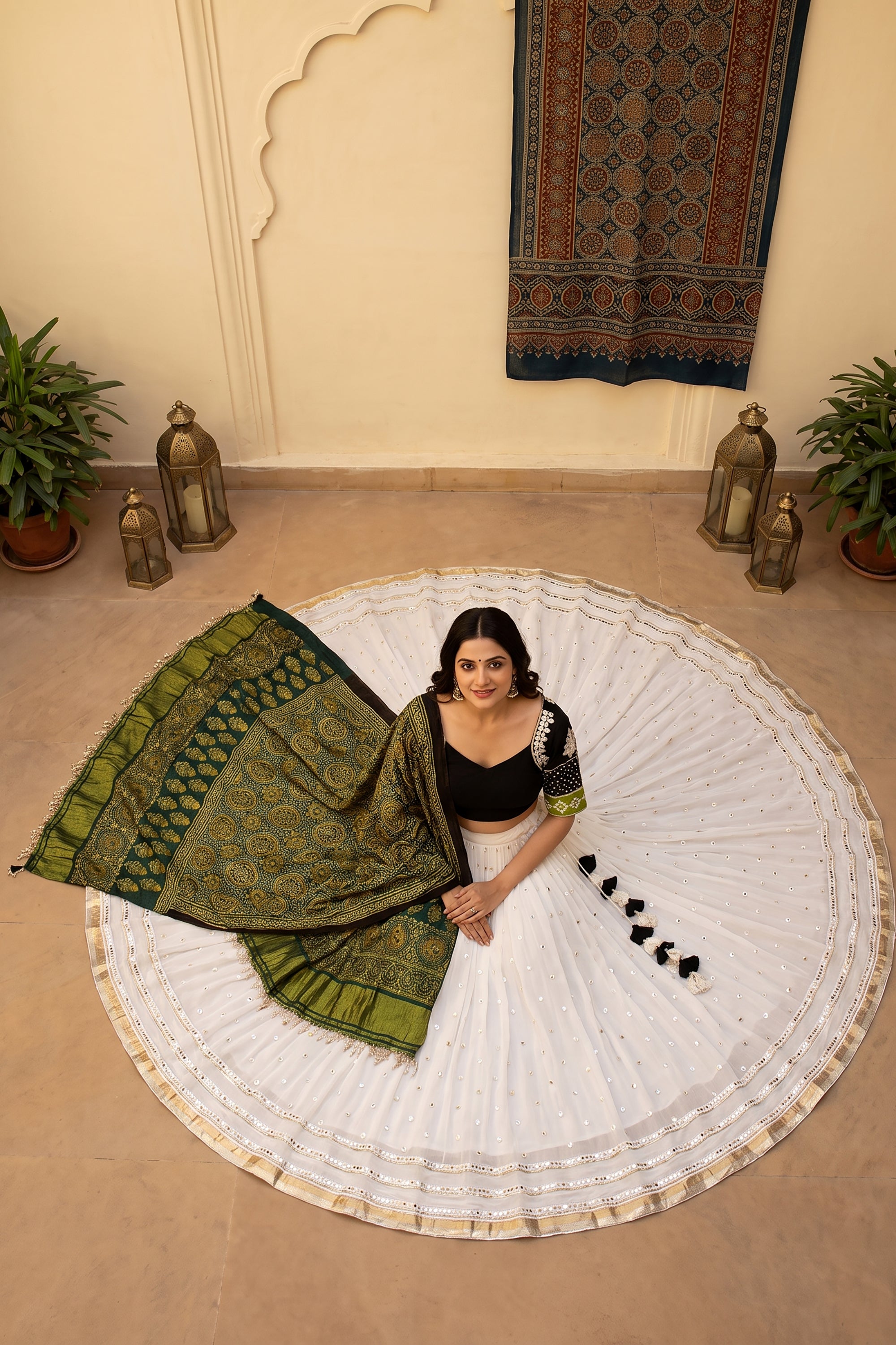 A woman smiling and twirling in a white flared georgette chaniya choli with subtle mirror work at the hem. The outfit is paired with a black V-neck blouse featuring embroidered sleeves and a draped green Ajrakh print dupatta.