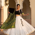 A woman smiling and twirling in a white flared georgette chaniya choli with subtle mirror work at the hem. The outfit is paired with a black V-neck blouse featuring embroidered sleeves and a draped green Ajrakh print dupatta.