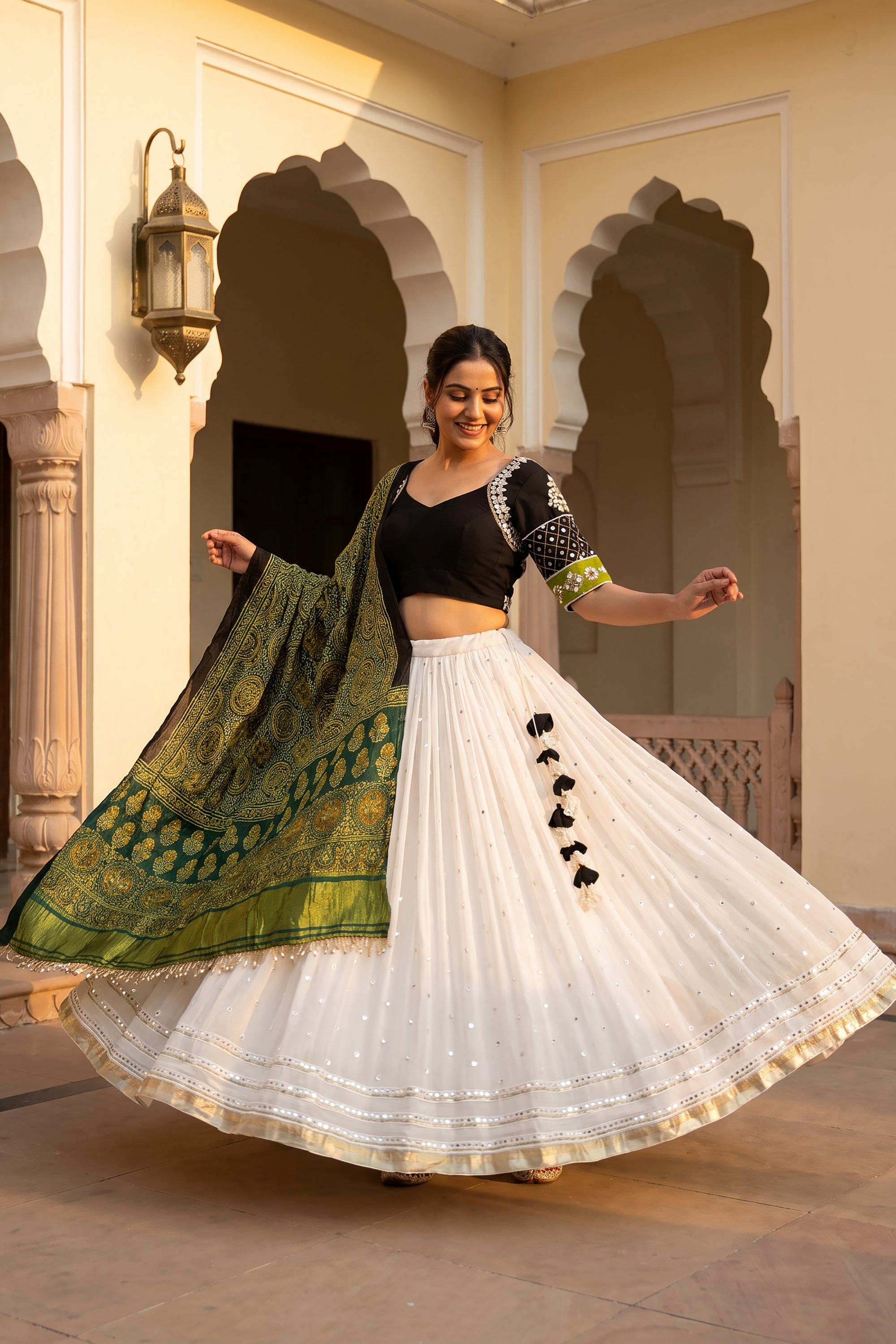 A woman smiling and twirling in a white flared georgette chaniya choli with subtle mirror work at the hem. The outfit is paired with a black V-neck blouse featuring embroidered sleeves and a draped green Ajrakh print dupatta.