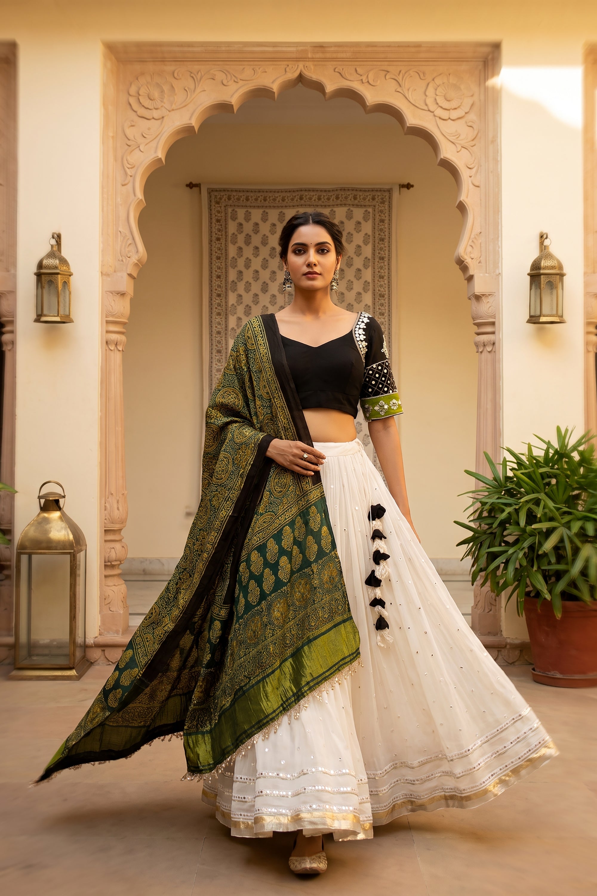 A woman smiling and twirling in a white flared georgette chaniya choli with subtle mirror work at the hem. The outfit is paired with a black V-neck blouse featuring embroidered sleeves and a draped green Ajrakh print dupatta.